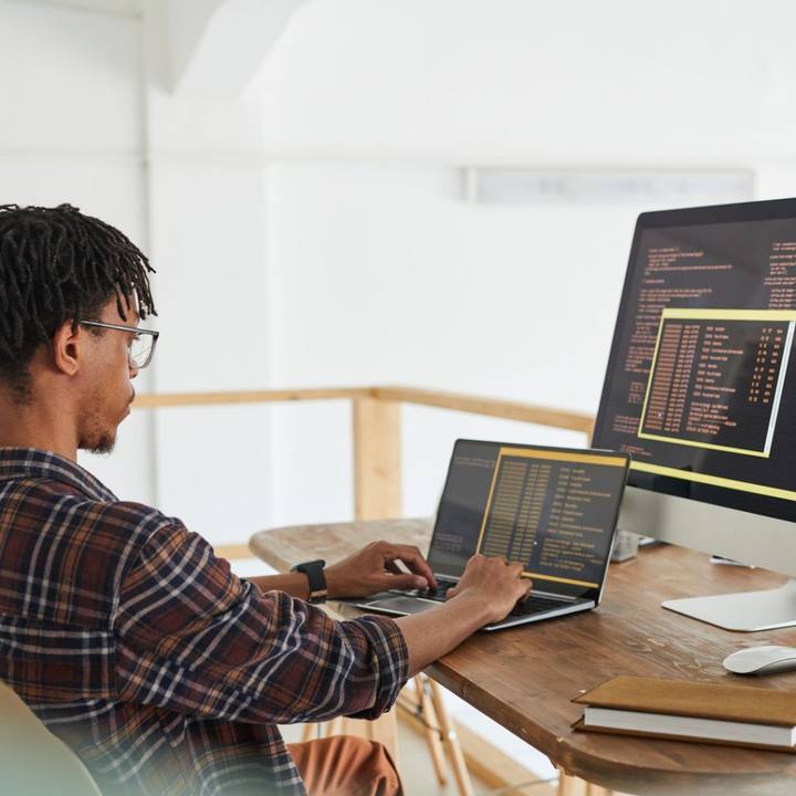 A man at a desk coding on a laptop beside a large monitor